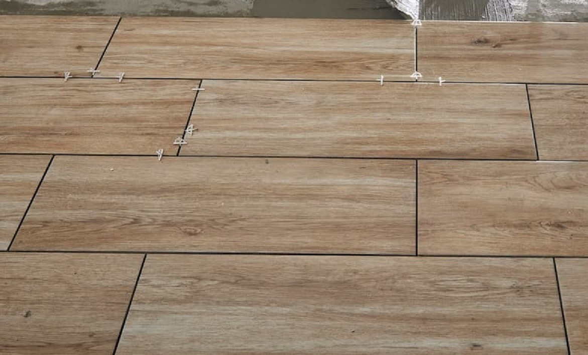 Układanie podłogi z płytek imitujących drewno, widoczne krzyżyki dystansowe i świeży klej na górze.