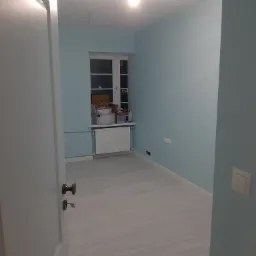 Jasne, błękitne wnętrze pokoju po remoncie, widoczne świeżo pomalowane ściany, białe okno z grzejnikiem i kartonami na parapecie, oraz fragment otwartych drzwi z brązową klamką.