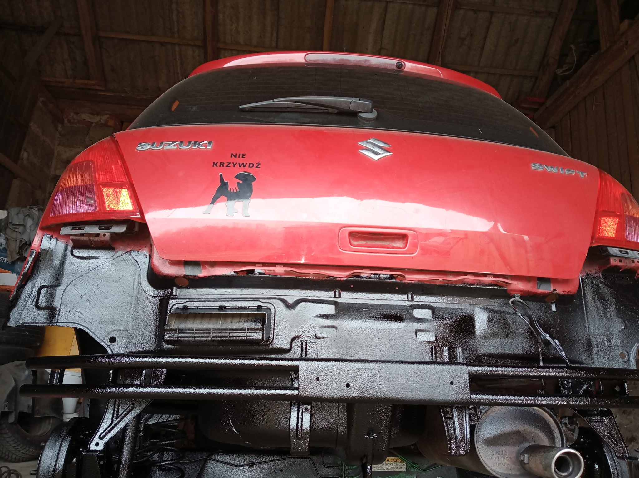 Czerwony Suzuki Swift z widocznym podwoziem po zabezpieczeniu antykorozyjnym, pokrytym świeżą, czarną powłoką ochronną. Widoczna naklejka z psem i napisem 'NIE KRZYWDŹ' na klapie bagażnika.
