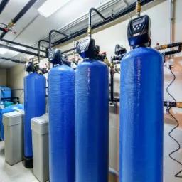 Rząd niebieskich zbiorników do uzdatniania wody z elektronicznymi wyświetlaczami, rurami i manometrami w pomieszczeniu technicznym z białymi kafelkami.