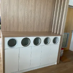 Biała szafka z pięcioma okrągłymi otworami na blacie, prawdopodobnie do segregacji śmieci, wkomponowana w drewnianą zabudowę.