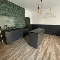 Nowoczesna, minimalistyczna kuchnia z czarnymi szafkami, zielonymi płytkami na ścianie i drewnopodobną podłogą, oświetlona dwoma wiszącymi lampami z żarówkami w stylu retro oraz napisem 'Fit cake...