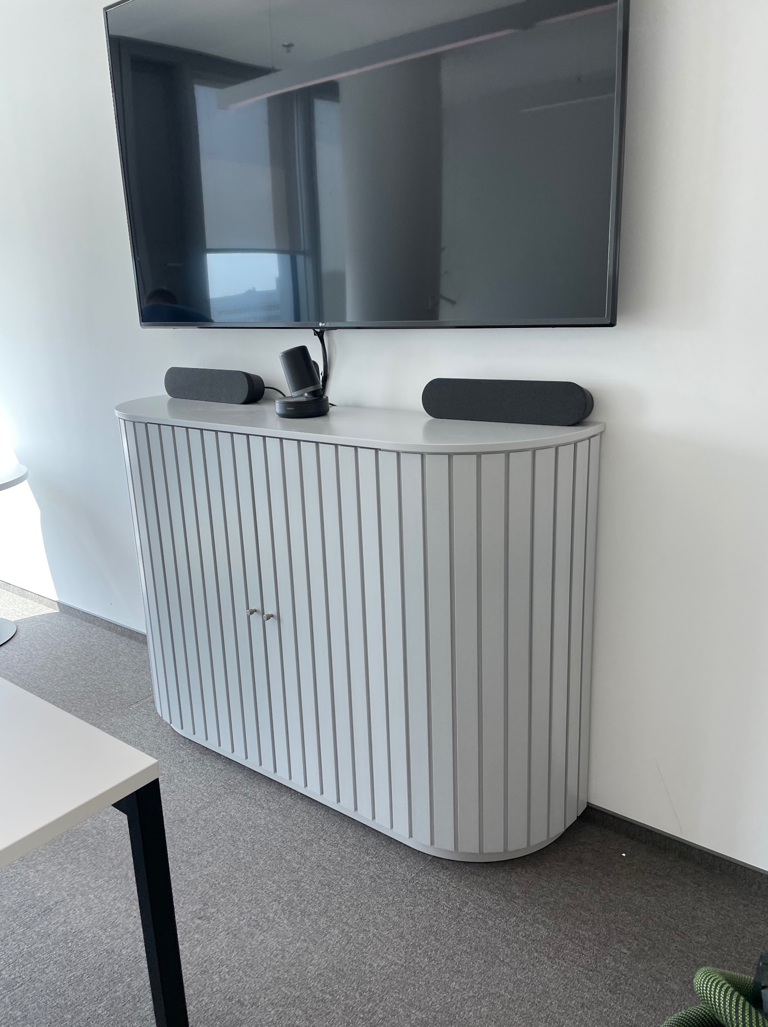 Szara szafka RTV z pionowymi żłobieniami, na niej głośniki i kamera do wideokonferencji, podwieszony telewizor na ścianie, minimalistyczne biuro.