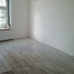 Pokój po remoncie z ułożoną podłogą z paneli imitujących drewno, widoczne ślady po czyszczeniu, białe ściany, okno z grzejnikiem i wisząca instalacja oświetleniowa.