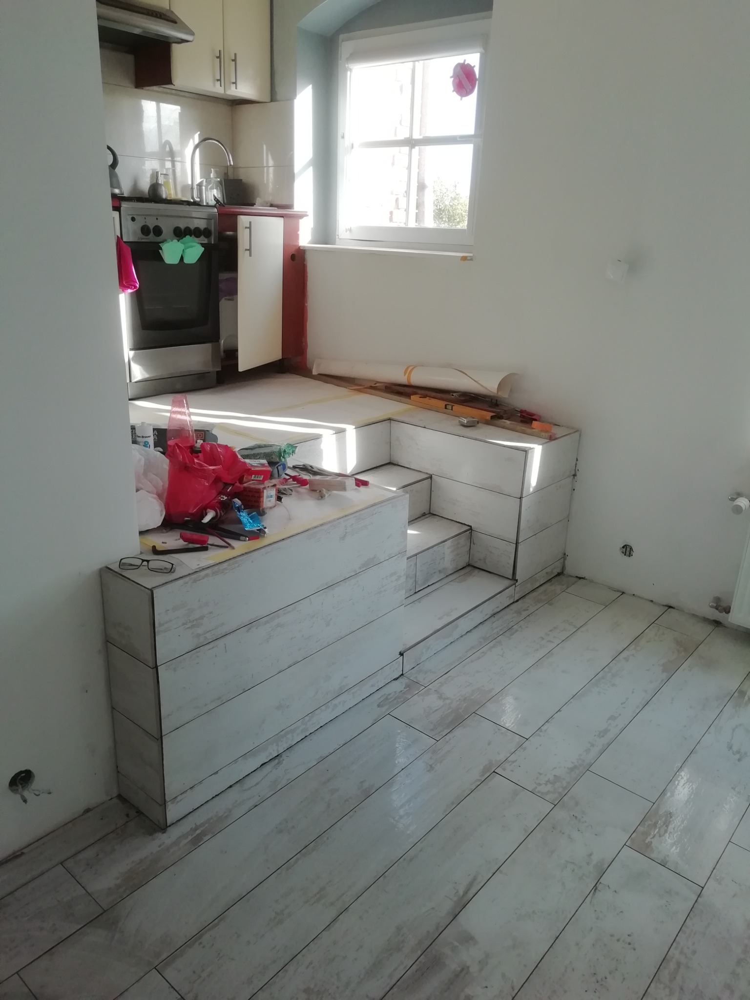 Wnętrze kuchni w trakcie remontu, widoczne ułożone płytki podłogowe imitujące drewno oraz konstrukcja z płytek tworząca podest z półkami, na której leżą narzędzia i akcesoria remontowe.