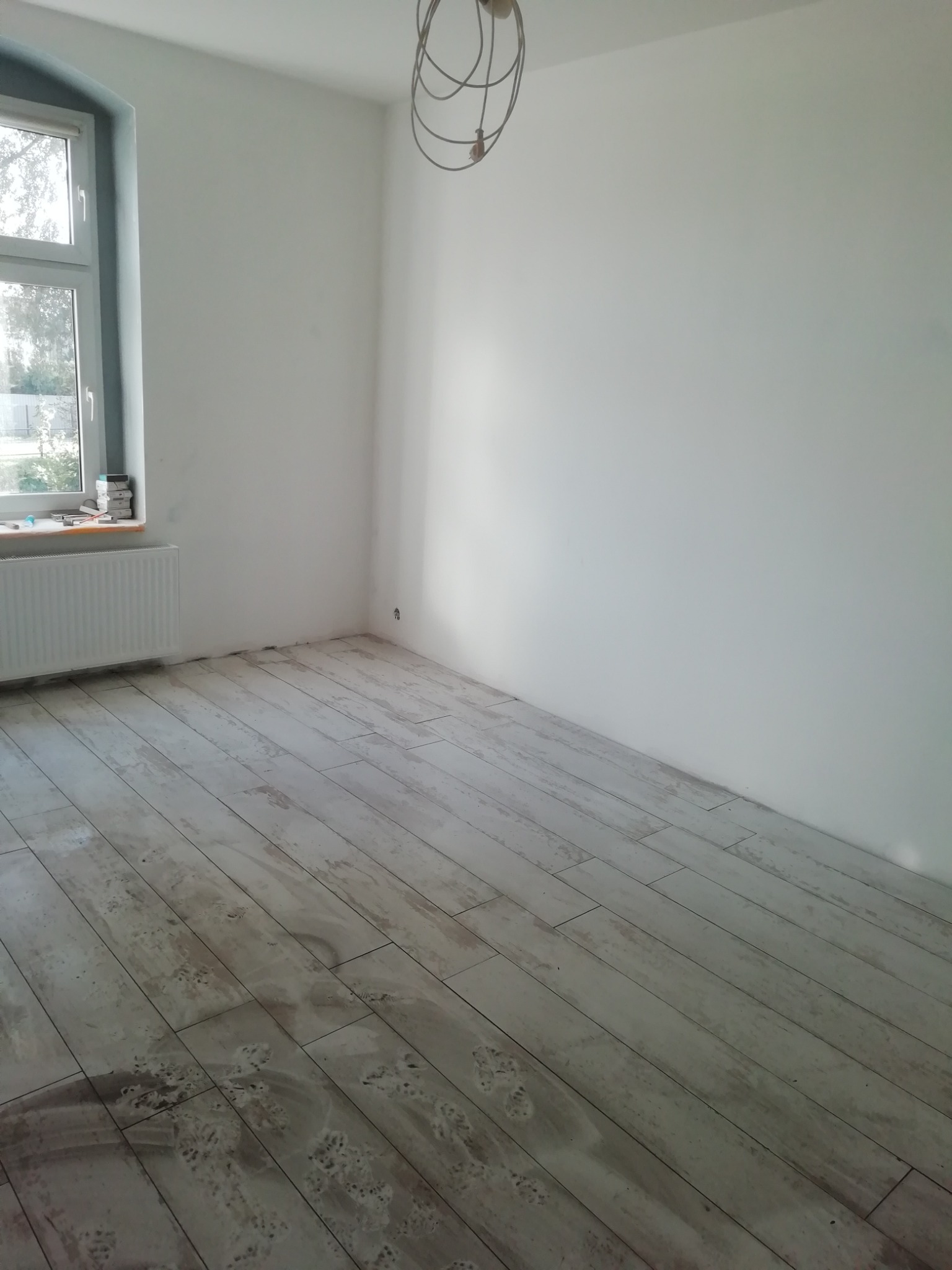 Pokój po remoncie z ułożoną podłogą z paneli imitujących drewno, widoczne ślady po czyszczeniu, białe ściany, okno z grzejnikiem i wisząca instalacja oświetleniowa.