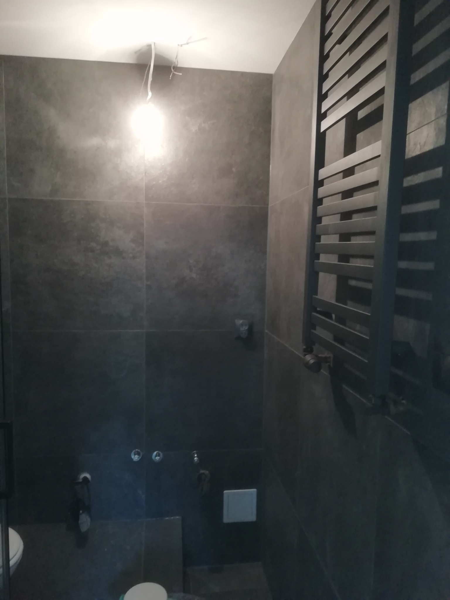 Ciemnoszara łazienka z dużymi płytkami, grzejnikiem drabinkowym i widocznymi instalacjami hydraulicznymi; surowy, nowoczesny styl w trakcie wykańczania.