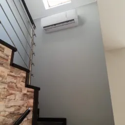 Biała jednostka klimatyzacji typu split zamontowana na szarej ścianie nad schodami z kamienną okładziną i metalową balustradą, widok z dołu, oświetlenie dzienne z okna dachowego.