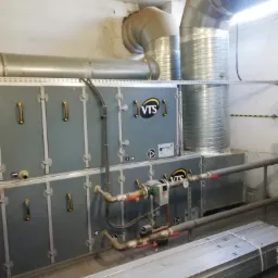 System wentylacji mechanicznej z rekuperacją VTS w kotłowni, widoczne rury wentylacyjne, zawory i elementy sterujące, na podłodze leżą profile montażowe.