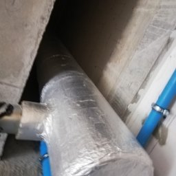 Ekomplex Klimatyzacje - Izolowany przewód wentylacyjny z odgałęzieniem, mocowany do sufitu, z widocznymi niebieskimi rurami i metalowymi opaskami zaciskowymi.