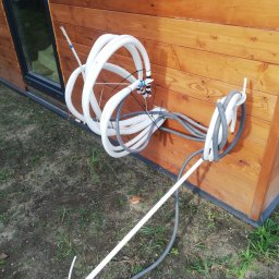 Ekomplex Klimatyzacje - Zewnętrzna instalacja przewodów klimatyzacyjnych i wentylacyjnych przy drewnianej ścianie budynku, widoczne białe rury izolowane i szare rury karbowane.