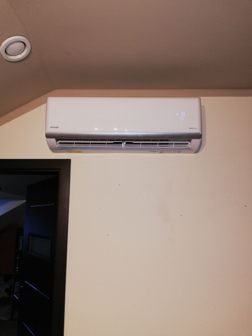 Biały klimatyzator marki Toyotomi zamontowany na jasnej ścianie wewnątrz pomieszczenia, widoczny fragment sufitu z okrągłym oświetleniem oraz fragment ciemnych drzwi po lewej stronie.