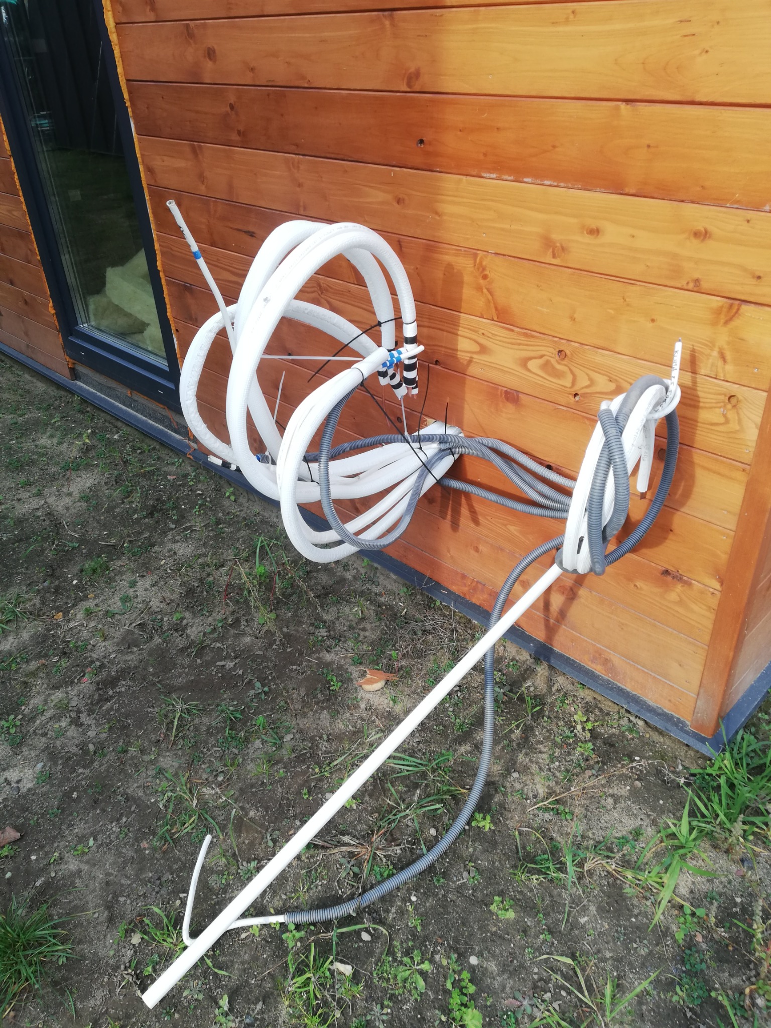 Zewnętrzna instalacja przewodów klimatyzacyjnych i wentylacyjnych przy drewnianej ścianie budynku, widoczne białe rury izolowane i szare rury karbowane.