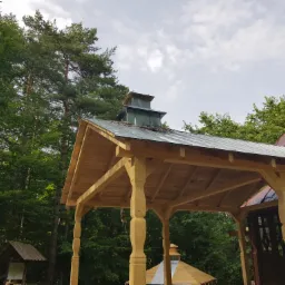 Wznoszenie drewnianej konstrukcji z metalowym zadaszeniem obok budynku sakralnego z czerwonym dachem, widok na las i pochmurne niebo.