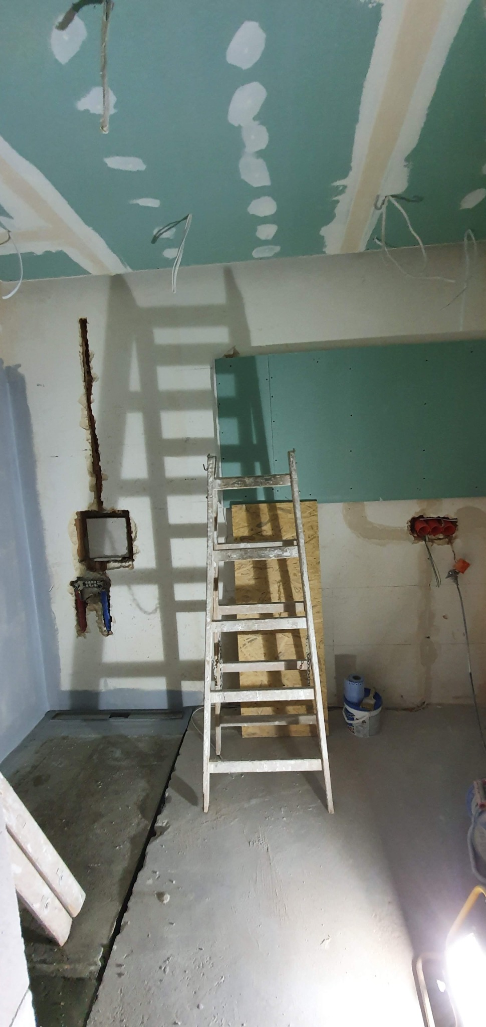 Wnętrze w trakcie prac przygotowawczych: surowy sufit z płyt kartonowo-gipsowych z widocznymi przewodami, ściany z przygotowanymi otworami na instalacje elektryczne i wodne, drabina oparta o ścianę...