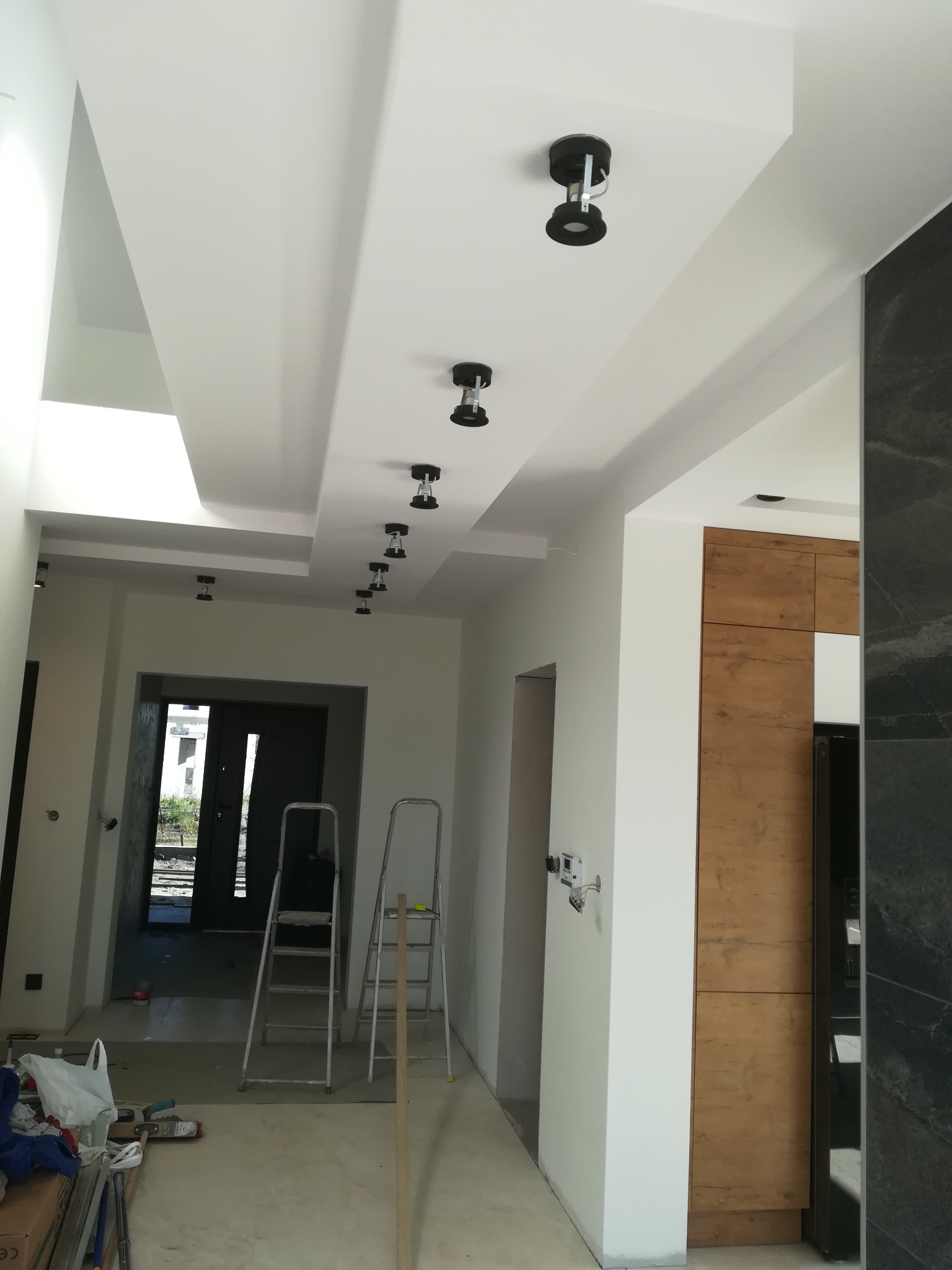Nowoczesne wnętrze w trakcie remontu z białymi ścianami, podwieszanym sufitem z zamontowanymi czarnymi reflektorami punktowymi, widoczne drabiny i narzędzia malarskie.