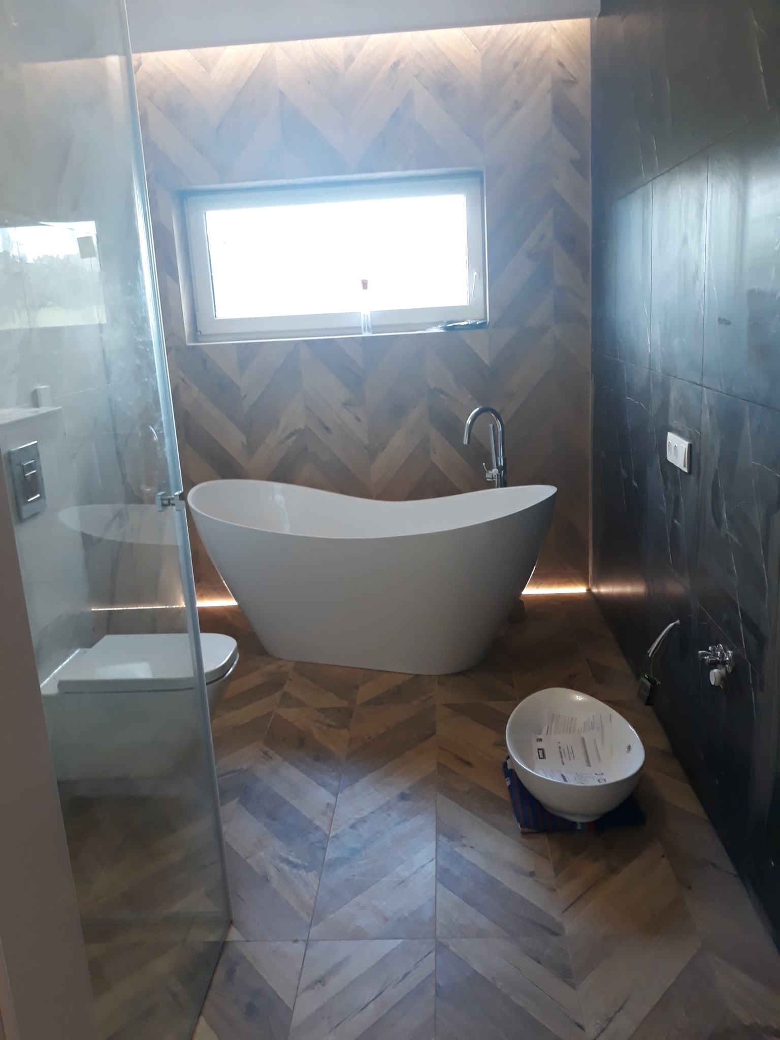 Nowoczesna łazienka z wolnostojącą wanną, drewnopodobną glazurą ułożoną w jodełkę na podłodze i ścianie za wanną, oraz ciemną glazurą imitującą marmur na pozostałych ścianach. Biała umywalka...