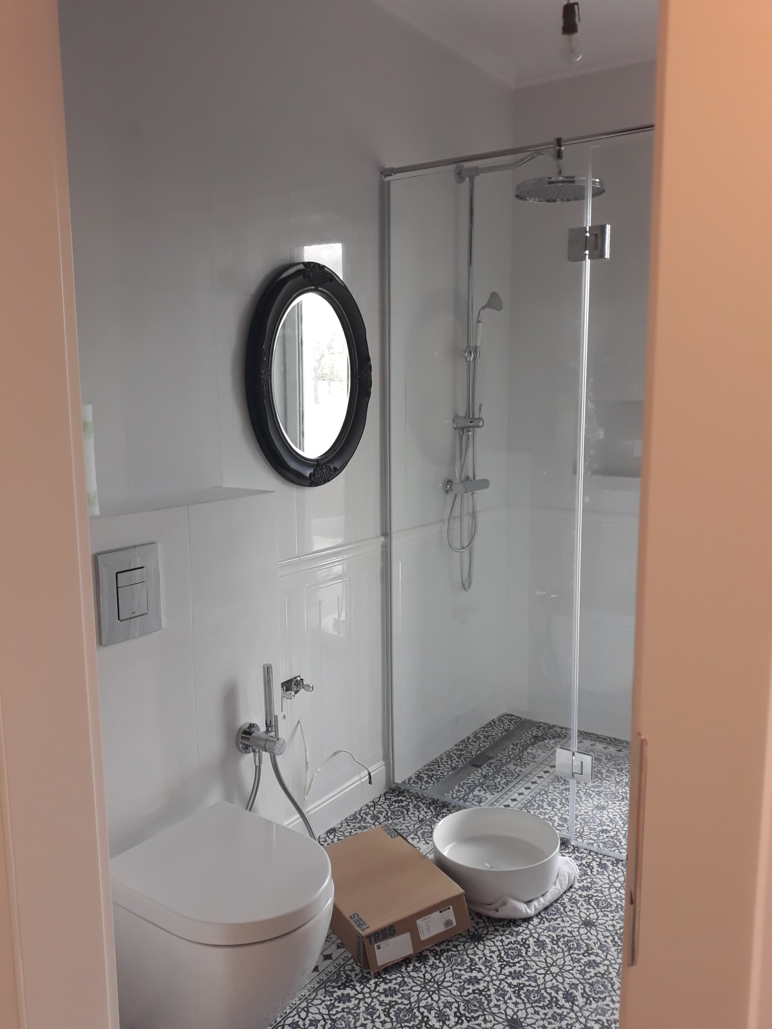Nowoczesna łazienka z kabiną prysznicową, toaletą z bidetem i okrągłym lustrem w czarnej ramie. Na podłodze wzorzyste płytki, obok pudełka i miska z wodą.