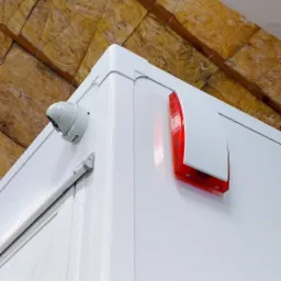 Kamera kopułkowa i sygnalizator alarmowy zamontowane na białej ścianie z izolacją stropu w tle.