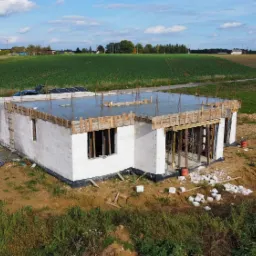 Surowy stan budynku jednorodzinnego z betonu komórkowego, z widocznym zbrojonym, zalanym stropem i drewnianym szalunkiem, na tle zielonych pól i błękitnego nieba.