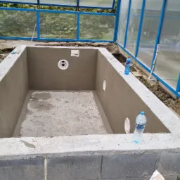 Surowy betonowy basen w trakcie budowy wewnątrz konstrukcji szklarni w Jastrzębiu-Zdroju, widoczne otwory instalacyjne i butelki wody na krawędzi.