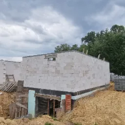 Wznoszenie ścian parterowego budynku z betonu komórkowego; widoczne zabezpieczenie fundamentów czarną izolacją, wykopane fundamenty pod kolejną część budynku, paleta bloczków betonowych obok.