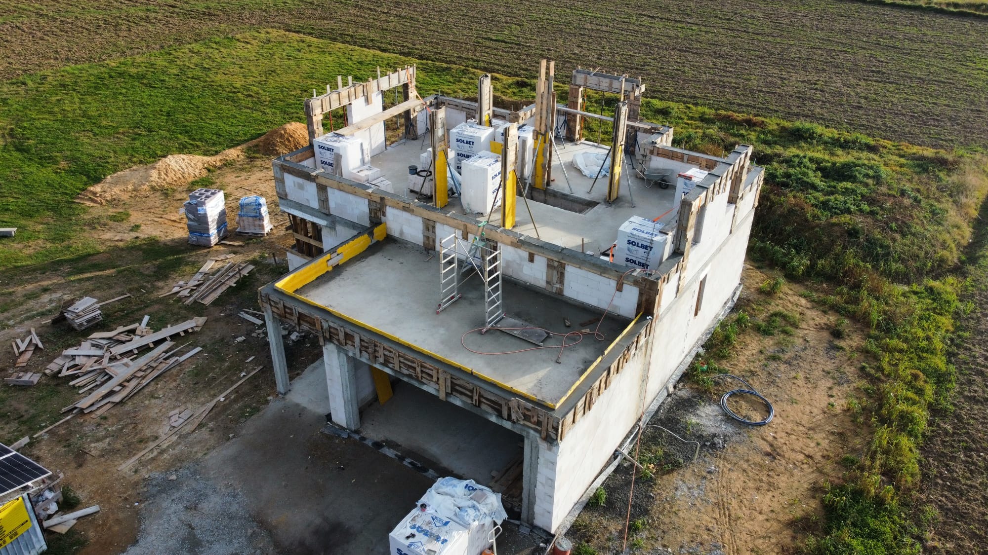 Wysoka perspektywa na budowę domu z bloczków Solbet, widoczne ściany parteru, szalunki stropu, drewniane stemple podtrzymujące, materiały budowlane na placu budowy i otaczające pole.