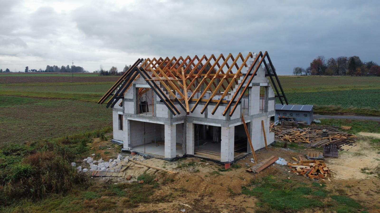 Niedokończony, dwukondygnacyjny budynek z białych bloczków, z częściowo zamontowaną drewnianą konstrukcją dachu, na tle pola i pochmurnego nieba.
