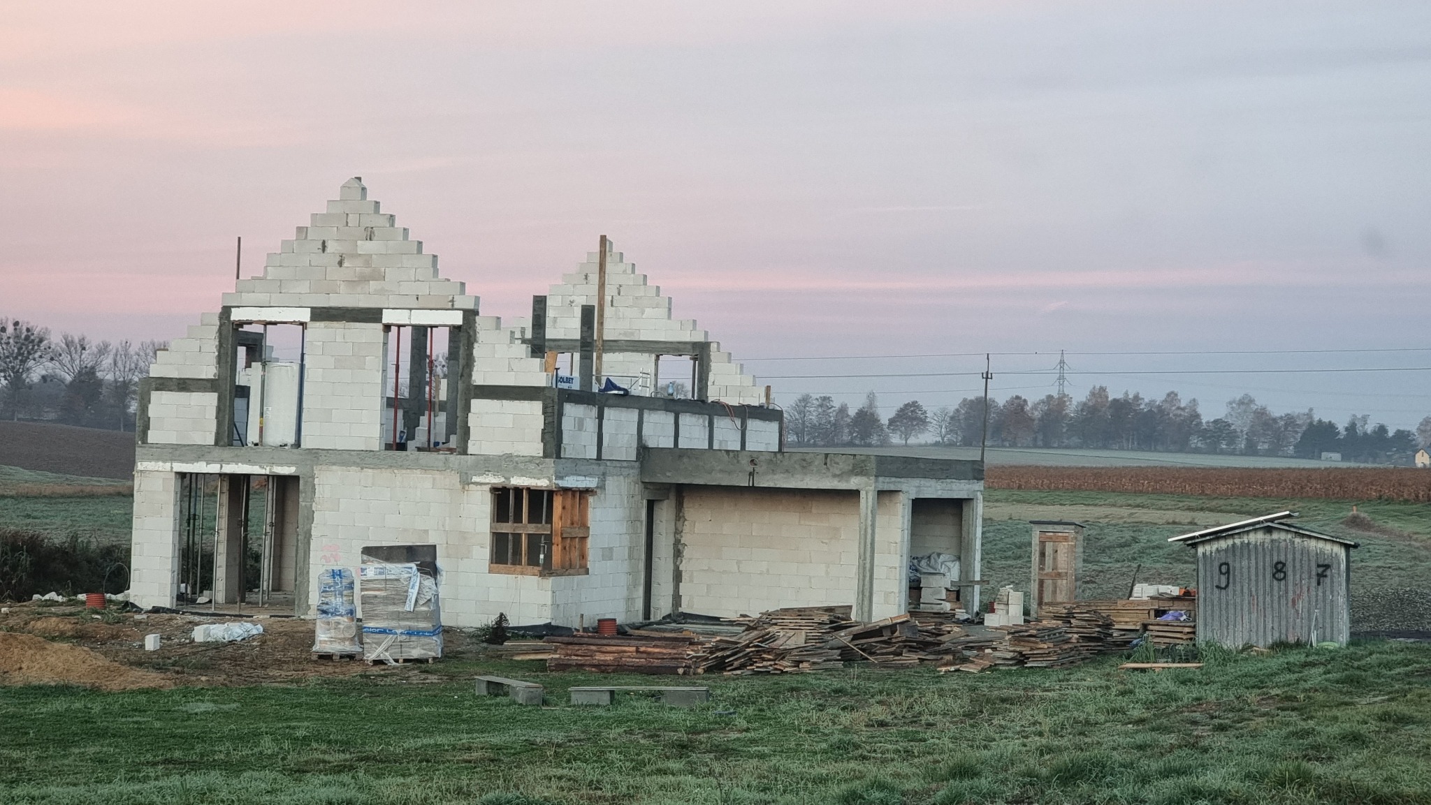 Wczesnym rankiem widok na budowę domu z białych bloczków, z charakterystycznym szczytem w kształcie piramidy, drewniane okno, palety z materiałami budowlanymi oraz szopa z numerem 987 w tle.