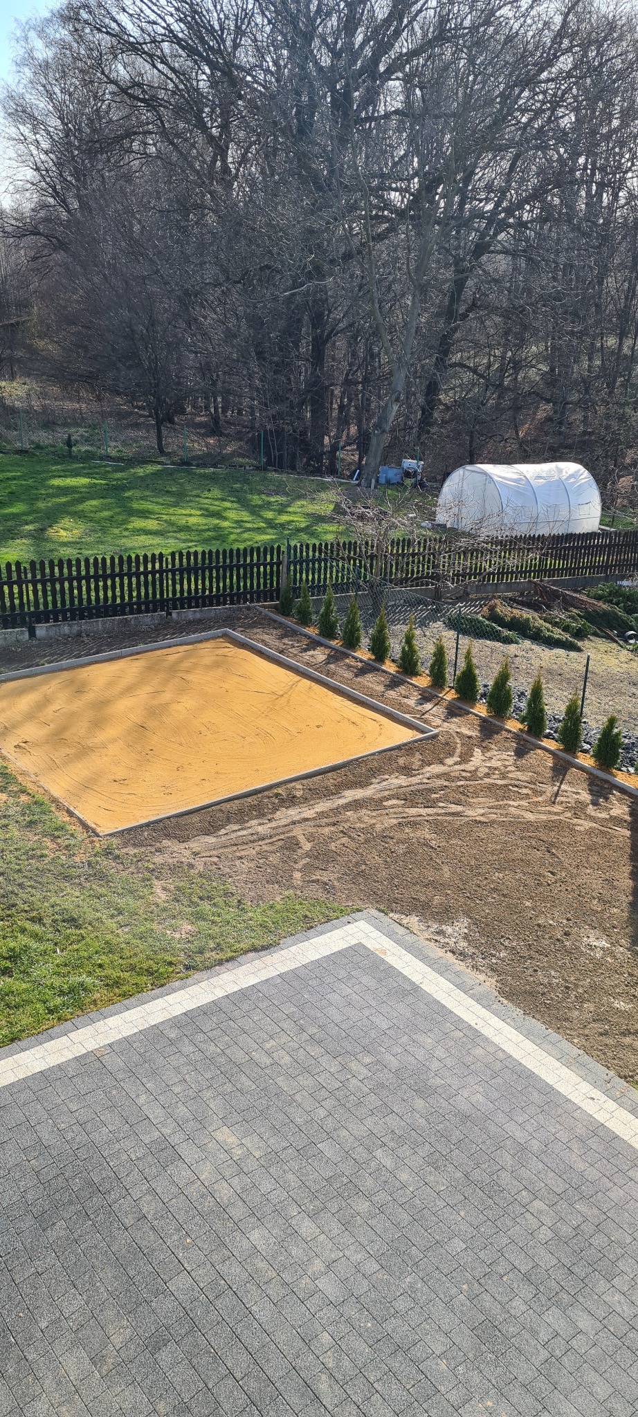 Przygotowany teren pod budowę, wyrównany piaskiem i obramowany betonowymi krawężnikami, z widoczną kostką brukową w tle, rzędem małych iglaków i tunelem foliowym na dalszym planie.