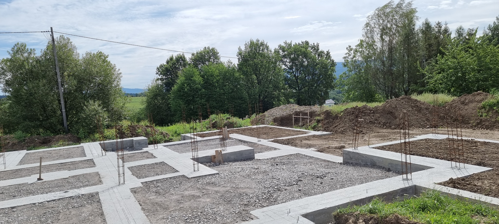Wylane betonowe fundamenty domu jednorodzinnego z wystającymi prętami zbrojeniowymi, widok z góry na tle zieleni i pagórkowatego krajobrazu.
