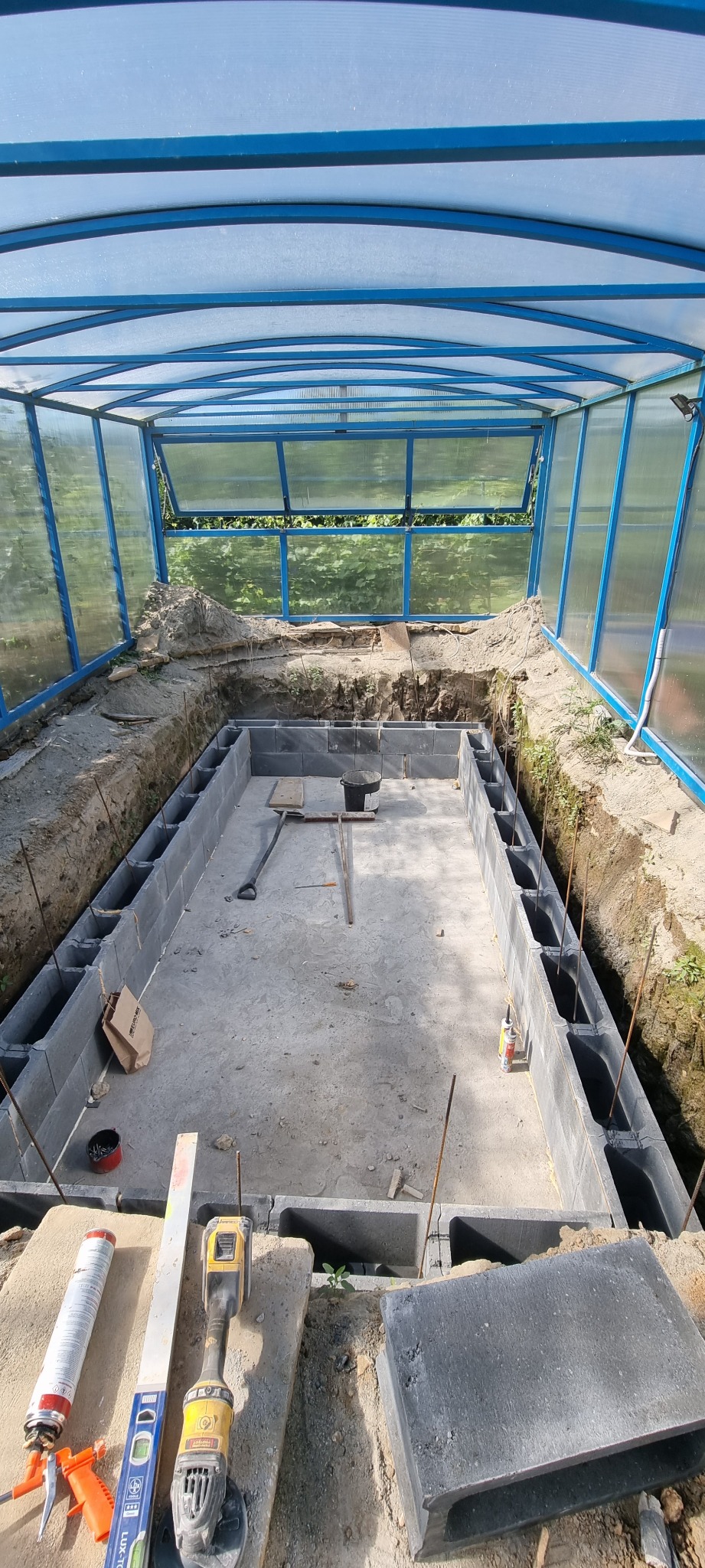 Wykop pod fundamenty basenu z szalunkiem z bloczków betonowych, widoczne narzędzia i materiały budowlane, zadaszenie z poliwęglanu.
