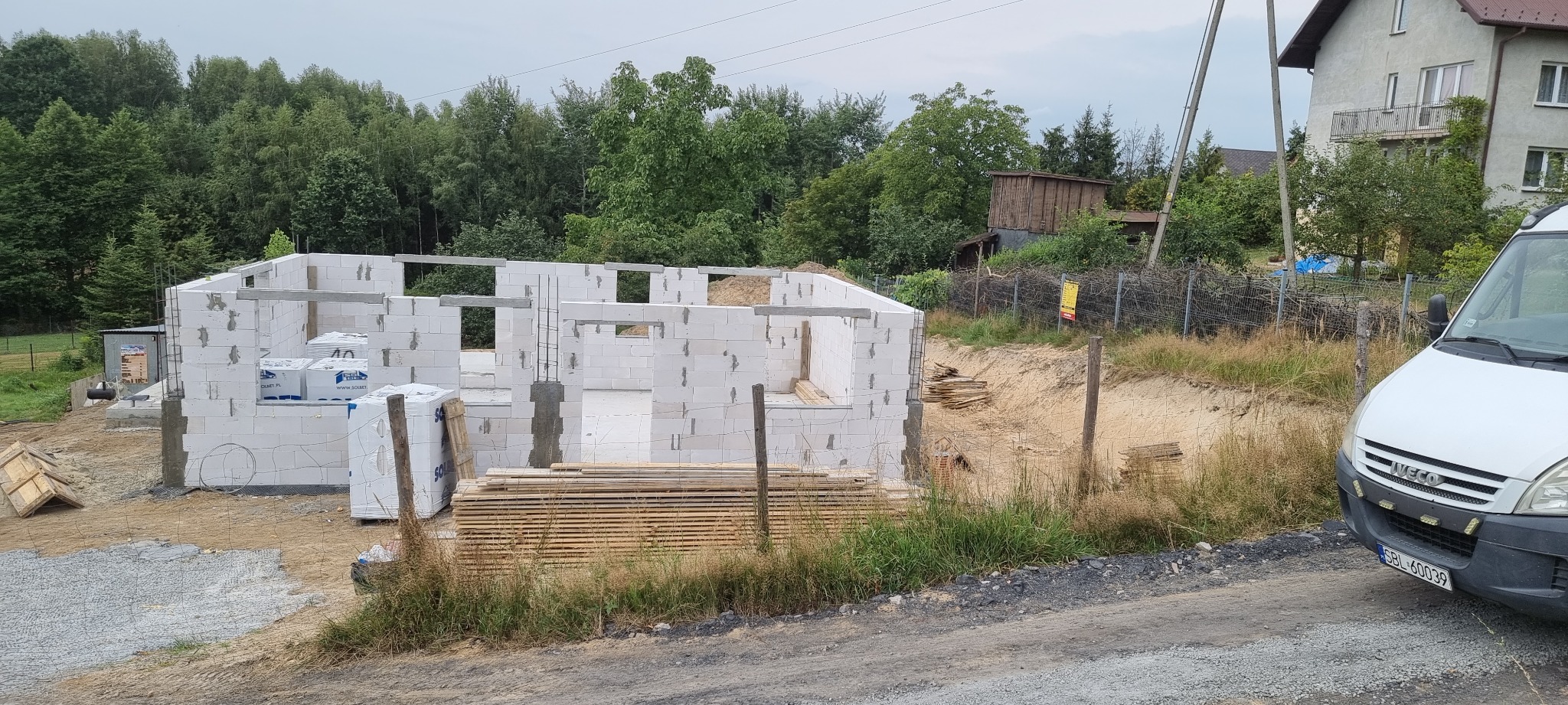 Wznoszenie ścian parterowego domu z białych bloczków, widoczne zbrojenie słupów, palety z materiałem budowlanym, w tle zalesione wzgórze i biały samochód dostawczy.