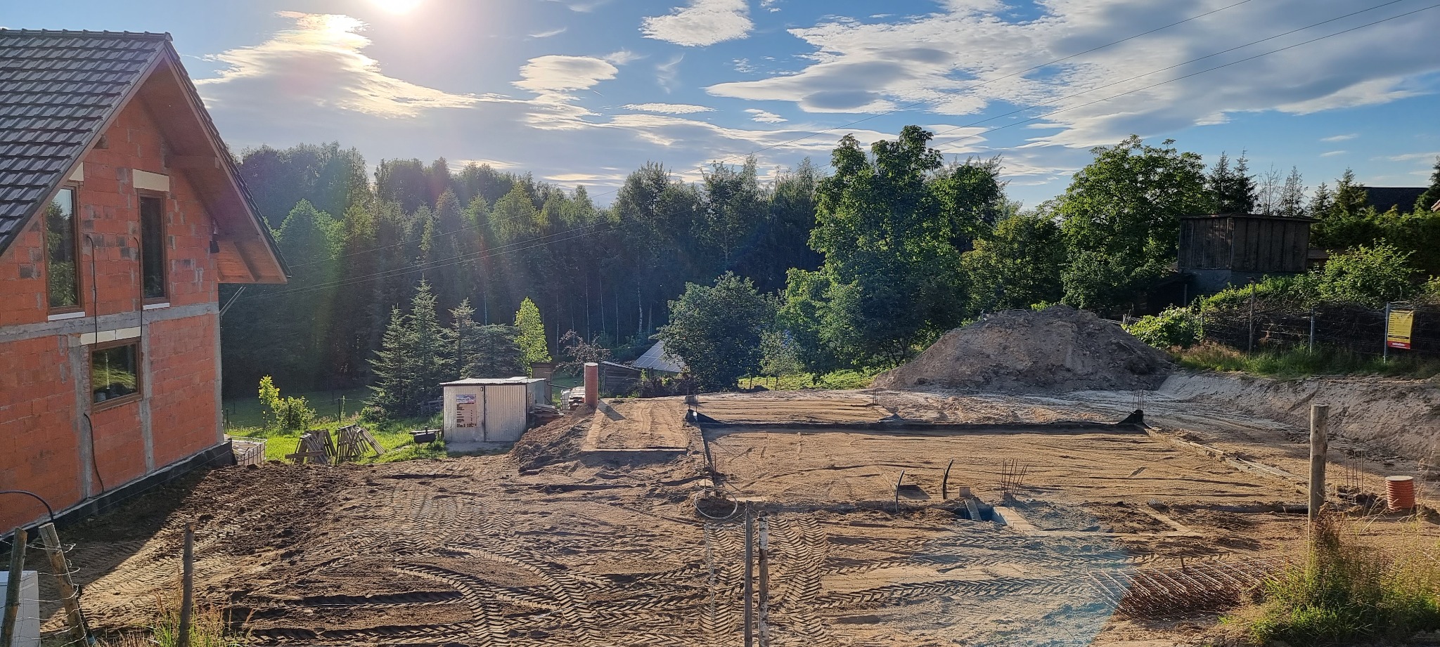 Budowa domu jednorodzinnego w stanie surowym otwartym: widoczne fundamenty, ściany z czerwonej cegły, teren przygotowany pod wylewkę, zalesione tło pod błękitnym niebem.