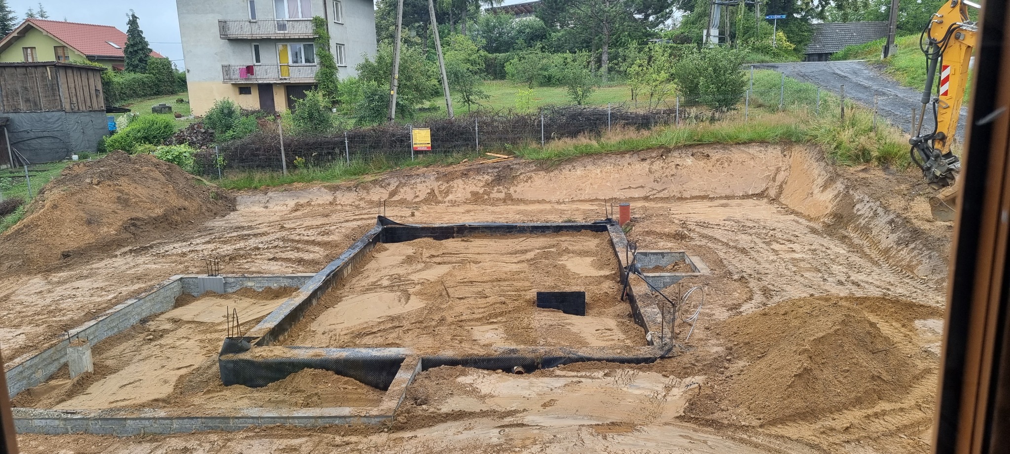 Wykop pod fundamenty z częściowo wylanymi ścianami fundamentowymi, widoczne zbrojenia, folia izolacyjna i pomarańczowa rura kanalizacyjna, na drugim planie wykopana ziemia i budynki mieszkalne.