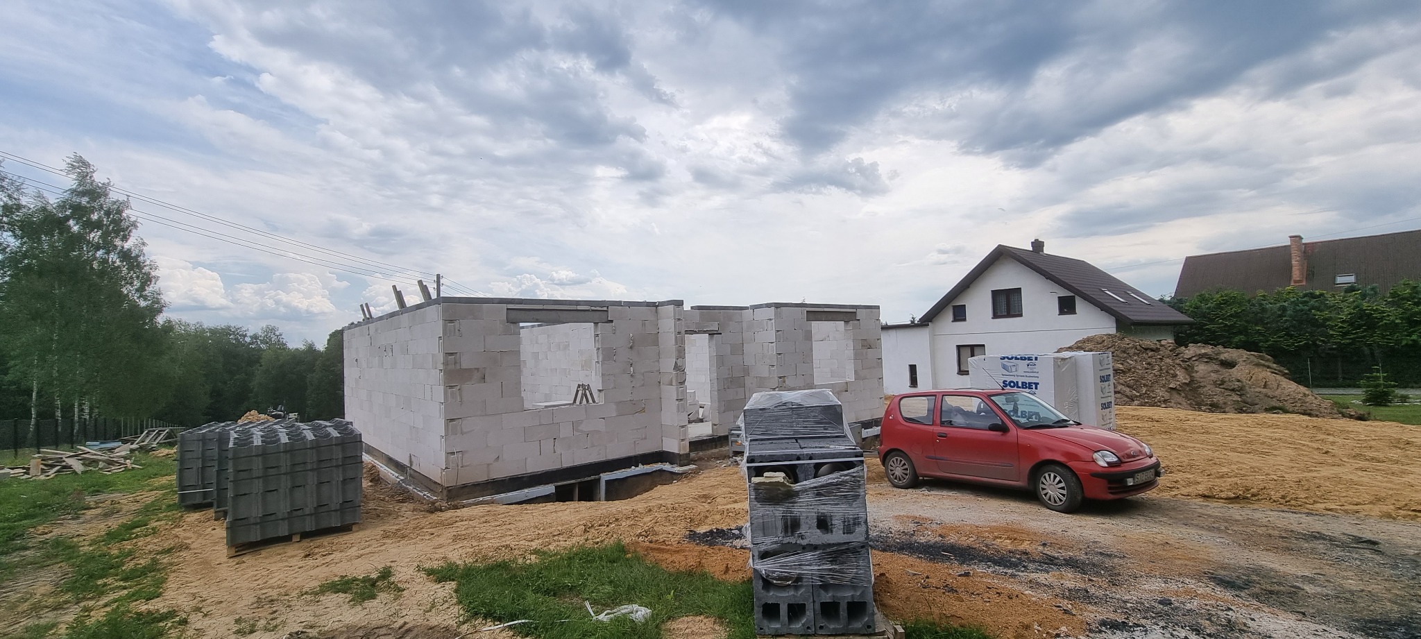 Wznoszenie ścian parterowego domu z bloczków betonu komórkowego, widoczna izolacja fundamentów, czerwony samochód osobowy na posesji, w tle zabudowa jednorodzinna.