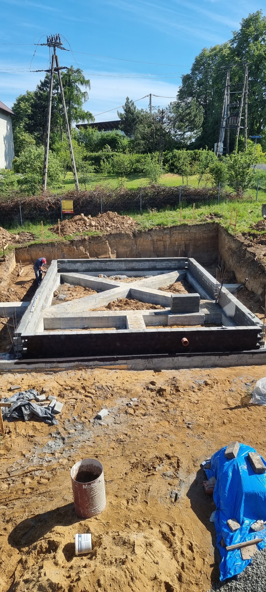 Wykop pod fundamenty domu z częściowo wylaną betonową konstrukcją, zabezpieczoną czarną izolacją poziomą; widoczny pracownik w oddali, teren budowy z elementami wyposażenia.