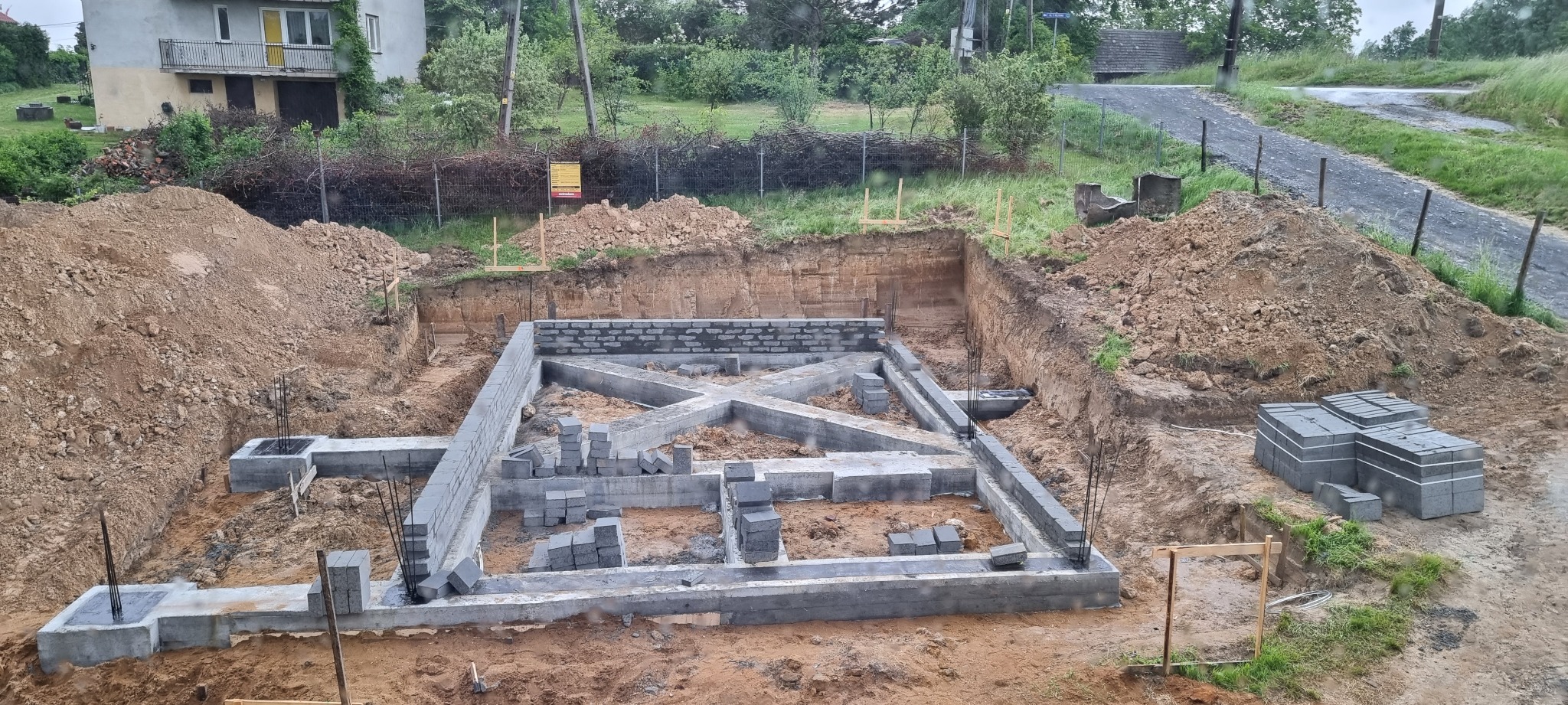 Wykop pod fundamenty z częściowo wzniesioną konstrukcją z bloczków betonowych, widoczne zbrojenia, stos bloczków obok wykopu, ziemia usypana wokół wykopu, w tle dom i droga.