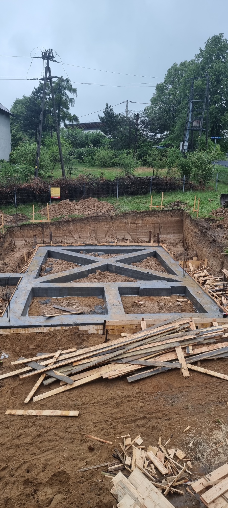 Wykop pod fundamenty z częściowo zalaną betonem konstrukcją w kształcie kratownicy, widoczne wystające pręty zbrojeniowe, deski szalunkowe i ziemia.