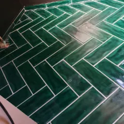 Ułożona zielona glazura w jodełkę na podłodze łazienki, widoczne fugi i fragment ściany w tle.