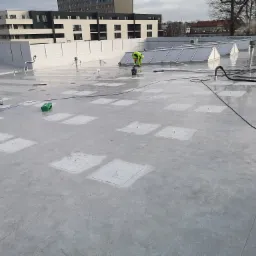 Prace na płaskim dachu pokrytym szarą membraną, robotnik w odblaskowej kamizelce używa szlifierki, widoczne skrzynki narzędziowe i kominki wentylacyjne.