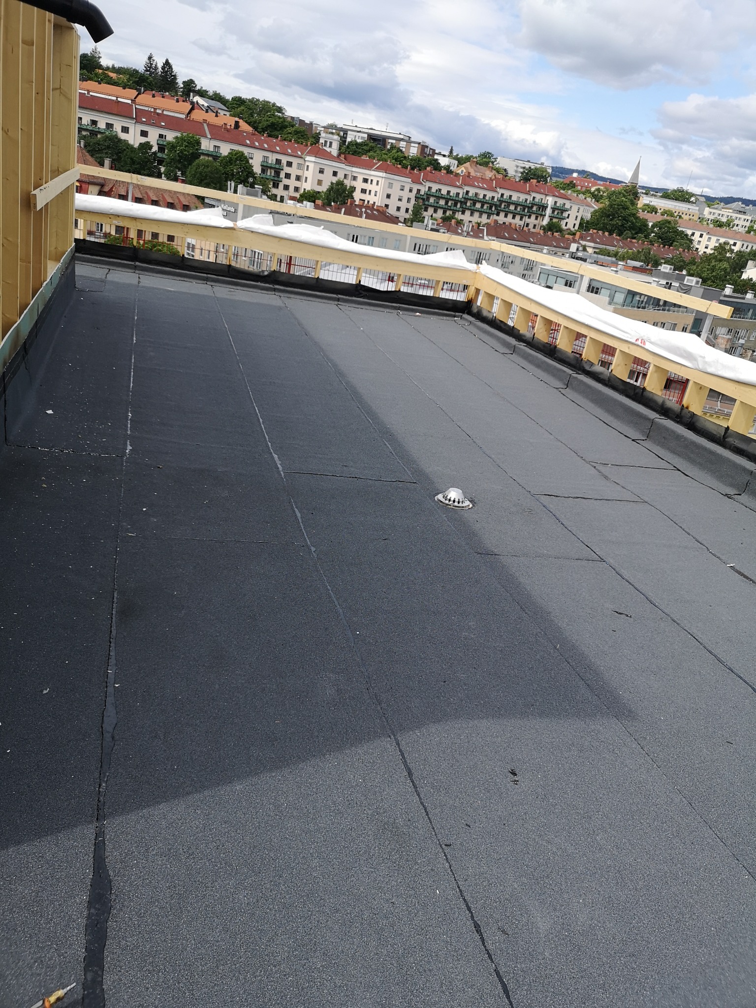 Wykończony, płaski dach pokryty papą termozgrzewalną w Gliwicach, z widocznym odpływem wody i fragmentem drewnianej konstrukcji attyki na tle zabudowy miejskiej.