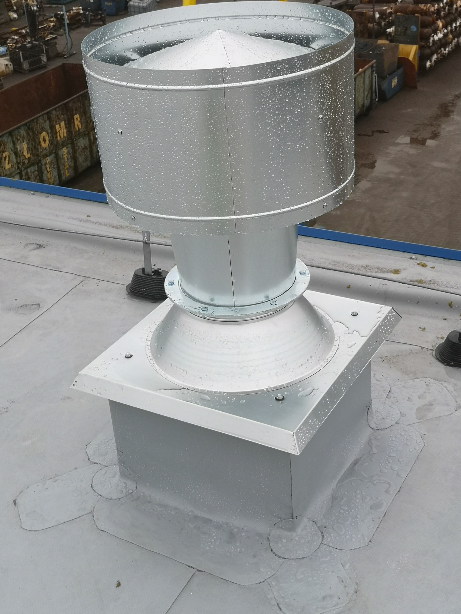 Metalowa nasada kominowa z kroplami wody na dachu przemysłowym z widocznym kontenerem na złom w tle.