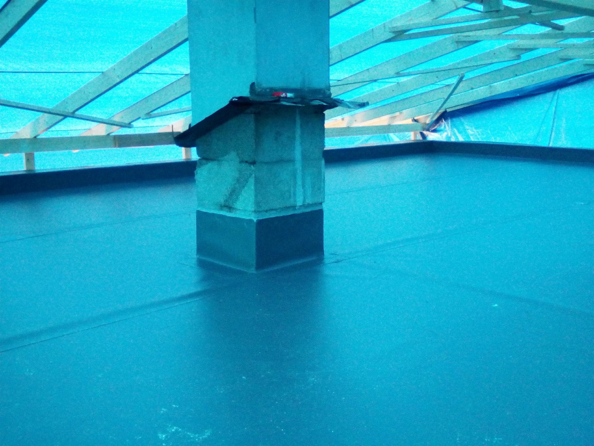 Płaski dach pokryty nową, jednolitą warstwą hydroizolacji w kolorze niebieskim, z wystającym kominem obłożonym cegłami i zabezpieczonym u podstawy czarnym pasem izolacyjnym; widoczne drewniane...