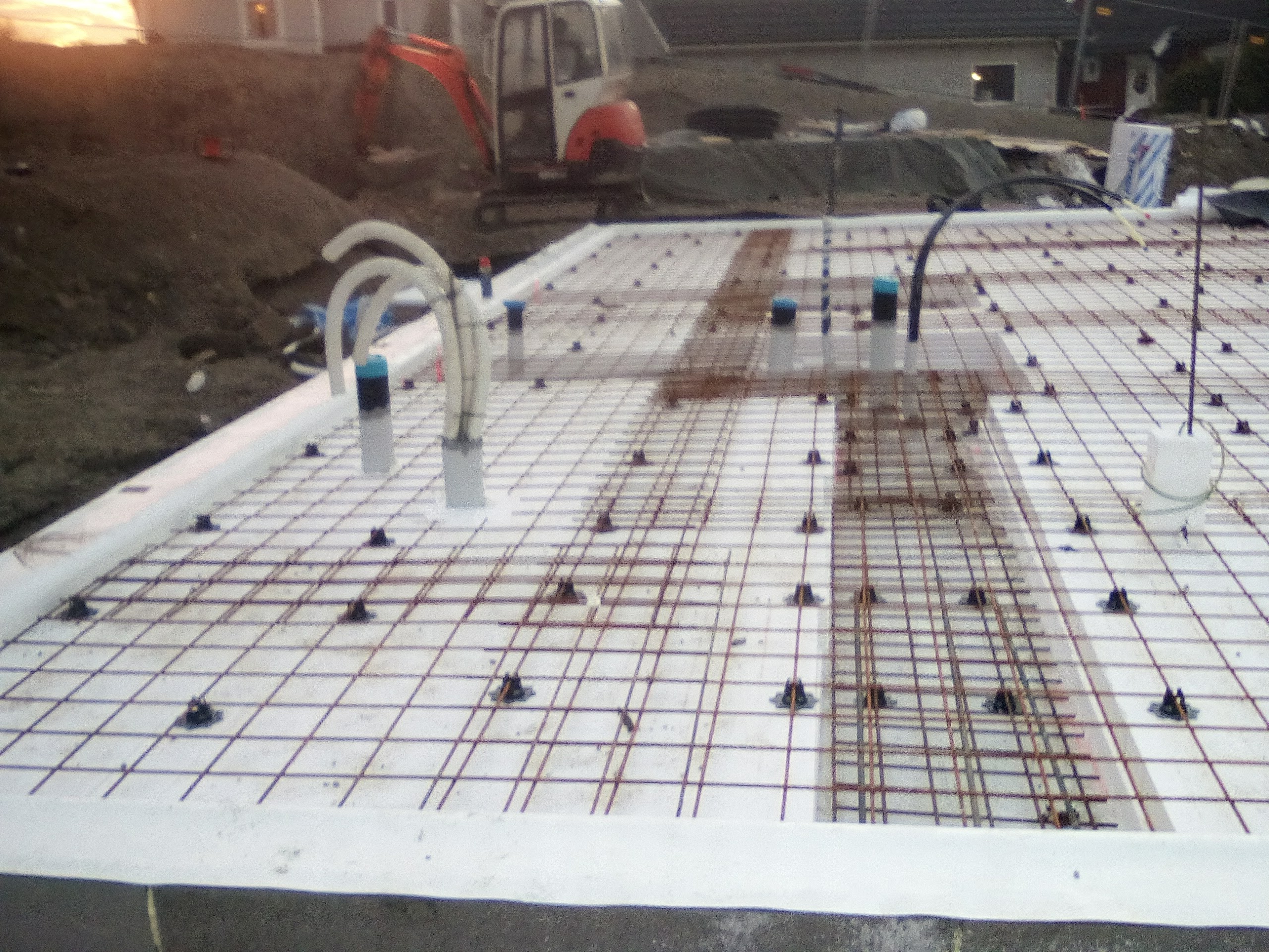 Przygotowanie płyty fundamentowej z izolacją termiczną i zbrojeniem stalowym, widoczne rury instalacyjne i minikoparka w tle.