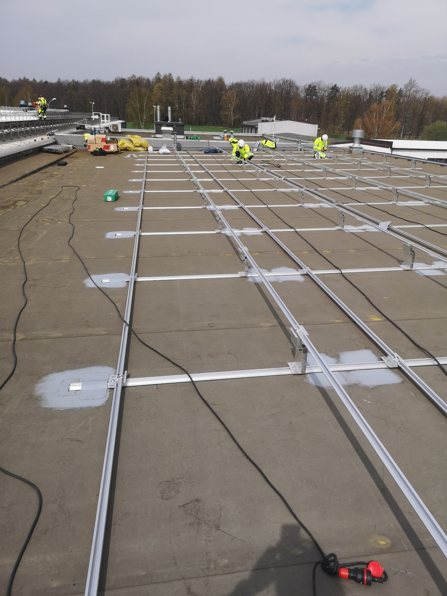 Montaż konstrukcji wsporczej pod panele fotowoltaiczne na płaskim dachu w Gliwicach. Widoczni pracownicy w odblaskowych ubraniach podczas pracy.