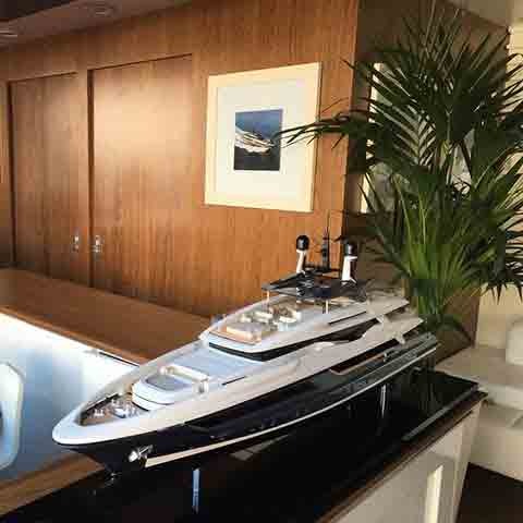 Model luksusowego jachtu motorowego na czarnym, błyszczącym stoliku, z drewnianą ścianą, obrazem i rośliną w tle.