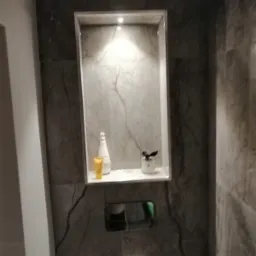 Wnęka w ścianie łazienki wyłożona szarymi płytkami imitującymi marmur, z oświetleniem punktowym i półką, na której stoją kosmetyki w białych i żółtych opakowaniach oraz mała roślina w doniczce...