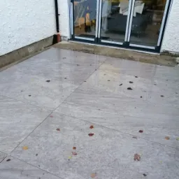 Szare płytki tarasowe z widocznymi fugami, pokryte opadłymi liśćmi, przylegające do ściany budynku z odpływem rynnowym i przeszklonymi drzwiami balkonowymi.