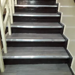 Schody wewnętrzne z szarymi panelami imitującymi drewno, zabezpieczone srebrnymi listwami antypoślizgowymi, przy jasnej ścianie i kremowej balustradzie.